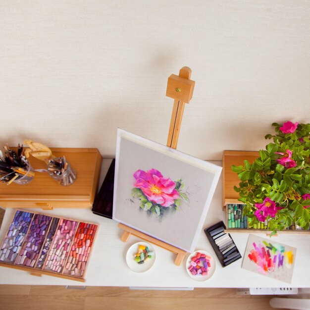 Houten schets ezel van een roze rozenbottel bloem staat op een tafel naast de materialen om te tekenen met penselen, potloden en droge pastel. concept van de werkplek van een vrouwelijke kunstenaar. copyspace