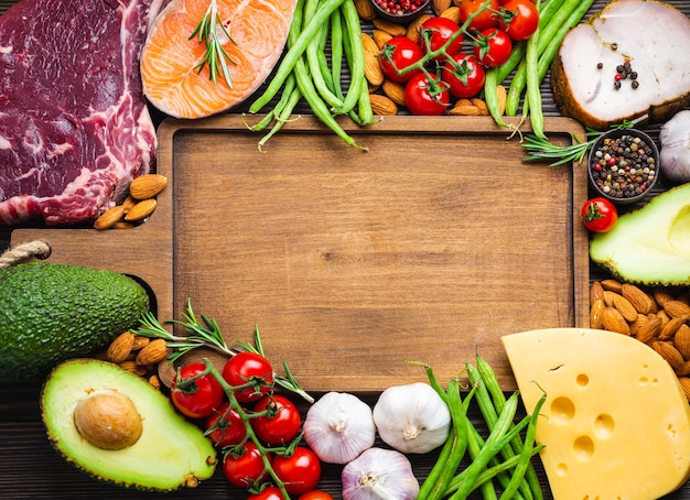 Houten snijplank en ketogene low carb-ingrediënten voor een gezond dieet voor gewichtsverlies, bovenaanzicht, kopieerruimte. Keto-voedingsmiddelen: vlees, vis, avocado, kaas, groenten, noten. Schoon eten, gezonde vetten