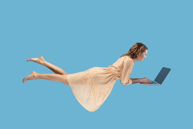 Hovering in air. Happy cheerful girl in yellow dress levitating with laptop, typing keyboard, reading good news message on computer while flying in mid-air. studio shot isolated on blue background