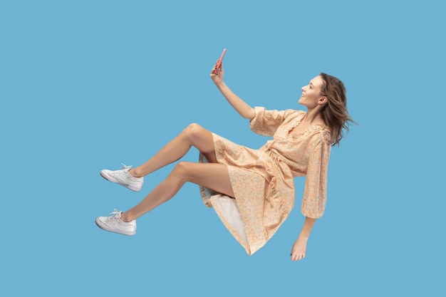 Hovering in air Smiling girl in yellow dress levitating with mobile phone reading message chatting happy joyful in social network online surfing web while flying indoor shot isolated on blue