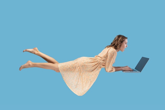 Hovering in air. surprised excited girl yellow dress levitating\
with laptop, typing keyboard, reading shocking news on laptop\
screen while flying in mid-air. studio shot isolated on blue\
background