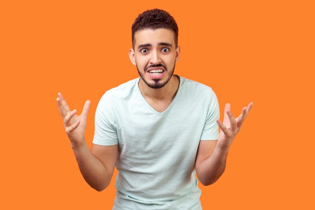 How could you. Portrait of annoyed frustrated brunette man with beard in white t-shirt standing with raised hands and indignant face asking why. indoor studio shot isolated on orange background