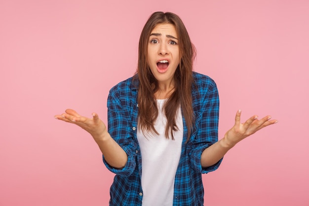 How could you Portrait of indignant girl in checkered shirt raising hands asking what why looking with dissatisfied disappointed expression making claims studio shot isolated on pink background