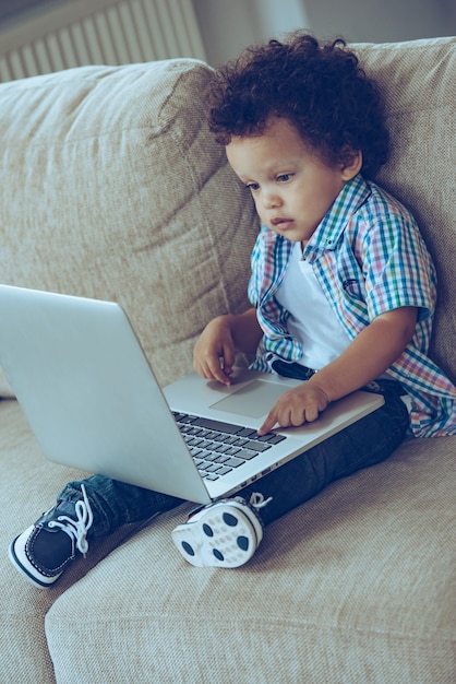 Come papà usa questa cosa? veduta dall'alto del piccolo bambino africano che usa il suo laptop mentre è seduto sul divano di casa