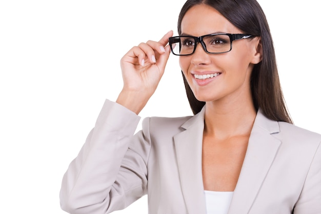 How may I help you? Beautiful young businesswoman in suit adjusting her eyeglasses and looking at camera while standing against white background