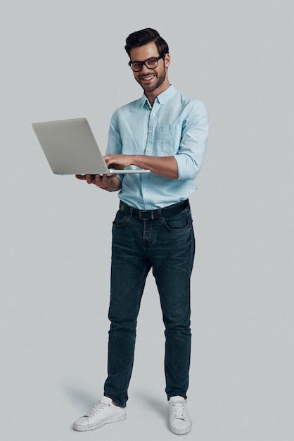 Come posso aiutarti? per tutta la lunghezza del giovane che usa il laptop e guarda la telecamera con un sorriso mentre si trova in piedi su uno sfondo grigio