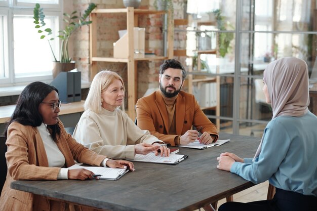 HR managers and head of company department sitting at table with resumes of Arabian job candidate and talking to her during meeting