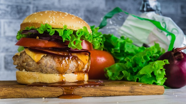 A huge juicy hamburger is on a chopping board with vegetables on a gray brick background