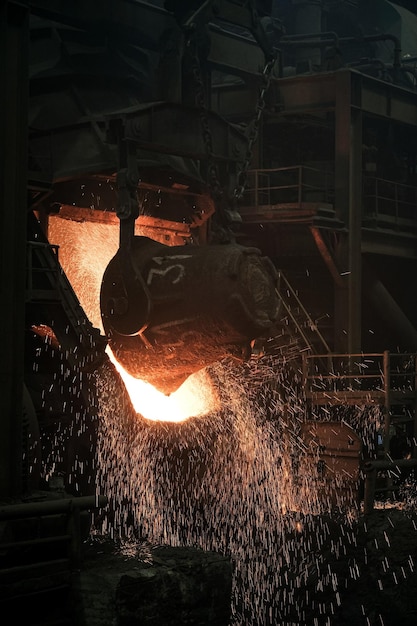 Огромный ковш наливает красное железо в печь на заводе, откуда летят искры. Крупным планом.