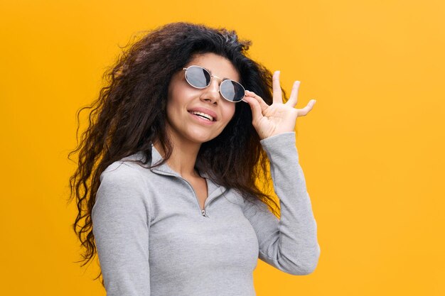 Huge sale promo concept portrait of happy pretty latin female\
with afro in sunglasses look at empty place touch eyewear glasses\
stay isolated over yellow background copy space free place\
banner