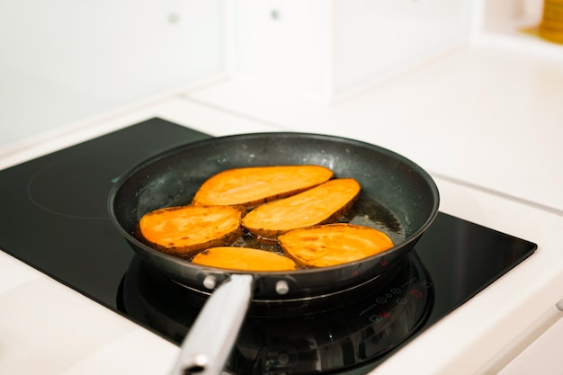 Huge sweet potatoes slices fries inside a frying pan. Food preparation. Roots. Vegetarian. Kitchen. Black induction hob. Meal. Dish. Yummy. Healthy food. Cooking