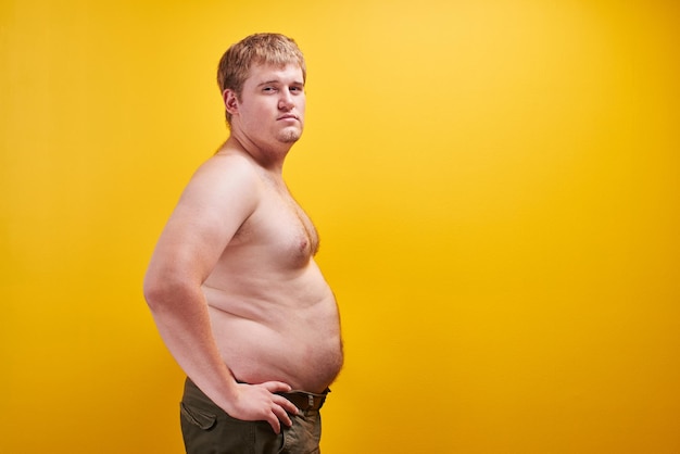 프로필에 큰 뚱뚱한 배와 벌거 벗은 몸을 가진 거대한 젊은 남자. 비만, 패스트푸드 및 정크 푸드, 스포츠, 지방흡입, 제품 또는 텍스트를 위한 여유 공간이 있는 체중 감소의 개념