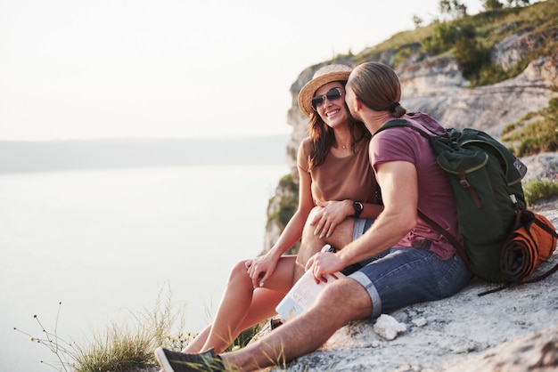 Hugging couple with backpack sitting on top of rock mountain enjoying view coast a river or lake. Traveling along mountains and coast, freedom and active lifestyle concept