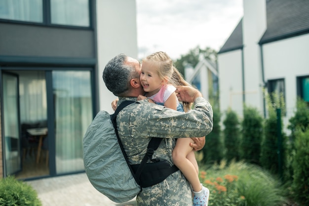 Abbracciando la sua tenera figlia. ufficiale militare che indossa l'uniforme e lo zaino che abbraccia la sua tenera figlia