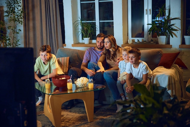 Photo hugging man and woman parents sitting on couch with their children and watching tv in the evening