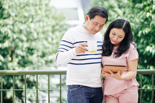 Hugging mature Asian husband and wife standing on balcony, drinking morning cofee and reading news on tablet computer