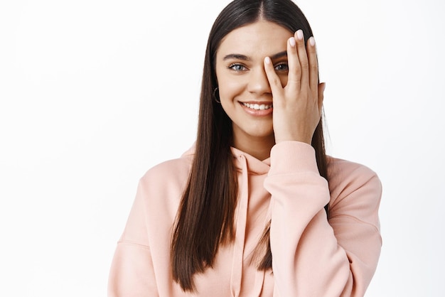 Huidverzorging en schoonheid Jong lachend brunette meisje dat de helft van het gezicht bedekt en door de vingers gluurt met een vrolijk gezicht in een casual hoodie tegen een witte achtergrond