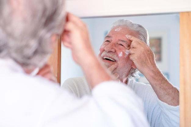 Huidverzorging knappe senior man die crème op zijn gezicht aanbrengt en naar zichzelf kijkt met een glimlach terwijl hij voor de spiegel staat