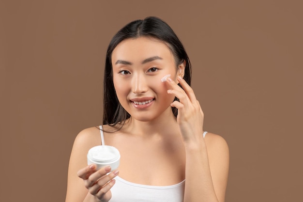 Huidverzorgingsroutine concept jonge aziatische vrouw die een pot met vochtinbrengende crème vasthoudt en aanbrengt