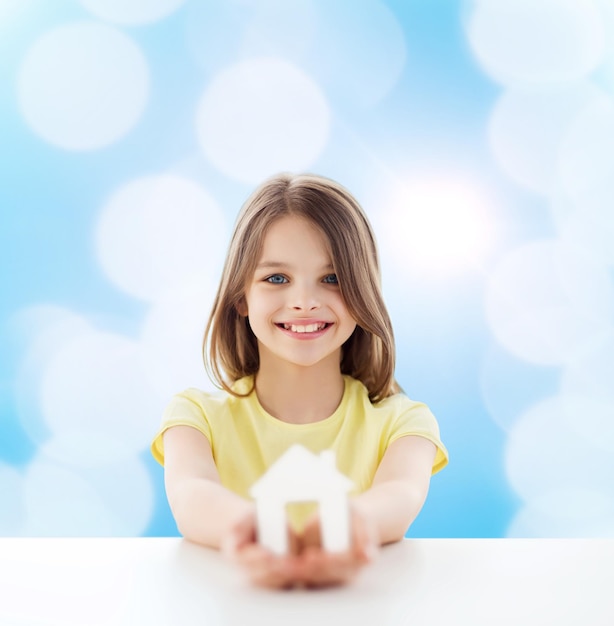 huis, onderwijs, geluk, jeugd en mensen concept - mooi klein meisje zittend aan tafel met witte huisknipsel over blauwe achtergrond