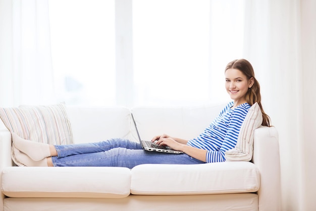 huis, technologie en internet concept - glimlachend tienermeisje liggend op de bank met laptop thuis