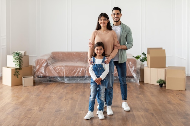 Huis verhuizen, renovatie en reparatie Concept. Volledige lichaamslengte van een vrolijk jong gezin van drie mensen die in een nieuw leeg appartement staan, poseren en kijken naar de camera, knuffelen, gratis kopieerruimte