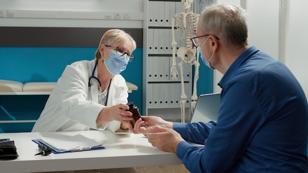 Huisarts en patiënt met gezichtsmasker praten over pillenfles in medisch kantoor. arts die voorschriftbehandeling en medicijnen geeft aan gepensioneerde man, om ziekte te genezen.