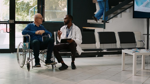 Huisarts in gesprek met senior man in faciliteitenlobby, medisch consult met patiënt met chronische handicap. Rolstoelgebruiker vraagt dokter om hulp en behandeling.