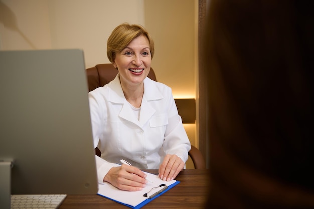 Huisarts in witte medische jas in gesprek met een vrouwelijke patiënt die haar raadpleegt en een formulier invult tijdens een consult tijdens een bezoek aan de kliniek