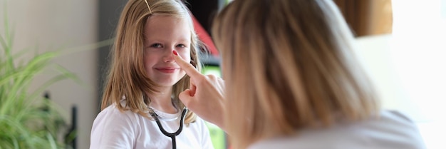 Foto huisarts raakt de neus aan van een schoolmeisje dat een stethoscoop vasthoudt. kinderarts speelt met haar jongen