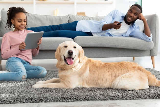 Huiselijk Leven Concept. Vrolijke zwarte familie die thuis rust, meisje zittend op vloertapijt met behulp van digitale tablet, man liggend op de bank met afstandsbediening die tv aan het schakelen is, focus op hond