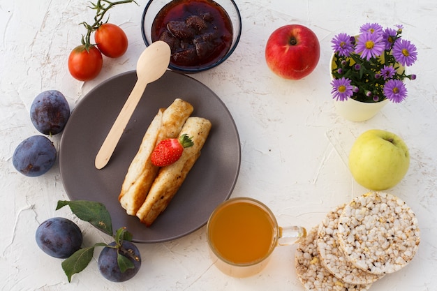 Huisgemaakte pannenkoeken gevuld met kwark, een aardbei op het bord, pruimen, een glazen kom met jam, een houten lepel, appels, gepofte rijstwafels, een kopje thee en bloemen