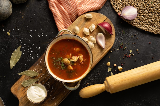 Huisgemaakte soep met dumplings, zure room, uien en kruiden. Een kom soep, een lepel, een handdoek, een deegroller en ingrediënten op een zwarte tafel. Bovenaanzicht.