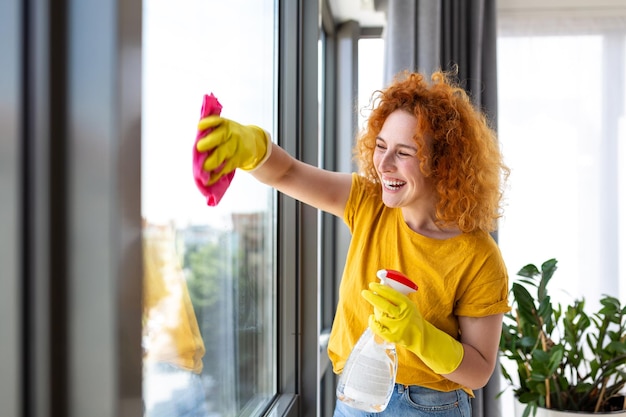 Huishoudelijk werk en huishoudelijk concept gelukkige vrouw in handschoenen die het raam thuis schoonmaakt met vod en reinigingsmiddel