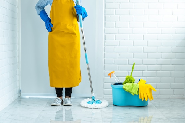 Huishouden en schoonmakend concept, Gelukkige jonge vrouw in blauwe rubberhandschoenen die stof afvegen die zwabber gebruiken terwijl thuis het schoonmaken op vloer