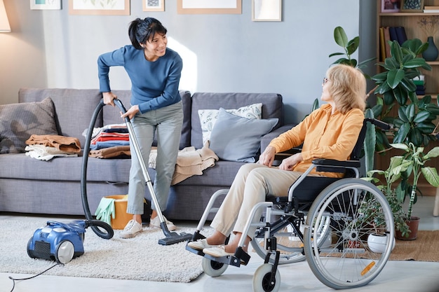 Huishoudster die stofzuiger gebruikt om het tapijt in de kamer schoon te maken, ze helpt senior vrouw die rolstoel gebruikt