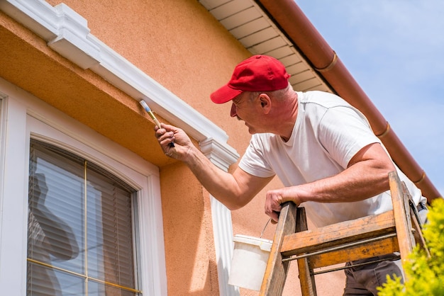 huisverzorging Een klusjesman op een ladder gebruikt witte verf voor de bovenramen van het huis. een man schildert