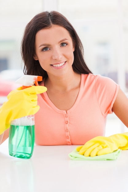 Huisvrouw op het werk. Jonge mooie vrouw in gele handschoenen die een vod en een spuitfles vasthoudt terwijl ze naar de camera glimlacht