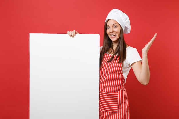 Huisvrouw vrouwelijke chef-kok bakker in gestreepte schort, wit t-shirt, toque chef-koks hoed geïsoleerd op rode achtergrond. Vrouw houdt groot wit leeg reclamebord voor promotionele inhoud. Bespotten kopie ruimte concept.