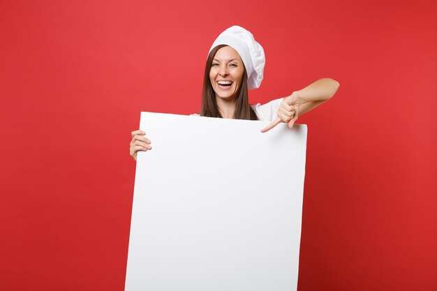 Huisvrouw vrouwelijke chef-kok bakker in gestreepte schort, wit t-shirt, toque chef-koks hoed geïsoleerd op rode achtergrond. Vrouw houdt groot wit leeg reclamebord voor promotionele inhoud. Bespotten kopie ruimte concept.