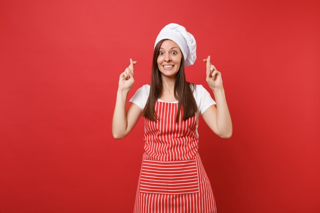 Huisvrouw vrouwelijke chef-kok of bakker in gestreepte schort, wit t-shirt, toque chef-koks hoed geïsoleerd op rode muur achtergrond. gelukkige vrouw die vingers gekruist houdt en wens doet. mock-up kopie ruimteconcept.