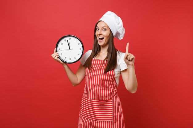 Huisvrouw vrouwelijke chef-kok of bakker in gestreepte schort, wit t-shirt, toque chef-koks hoed geïsoleerd op rode muur achtergrond. Glimlachende vrouw die de klok rond in de hand houdt, schiet op. Mock-up kopie ruimteconcept.