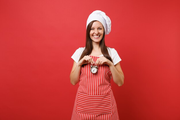 Huisvrouw vrouwelijke chef-kok of bakker in gestreepte schort, wit t-shirt, toque chef-koks hoed geïsoleerd op rode muur achtergrond. Glimlachende vrouw houdt in de hand retro wekker opschieten. Mock-up kopie ruimteconcept.