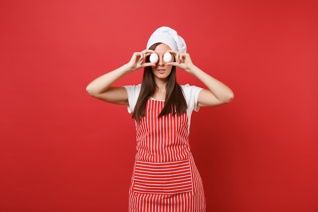 Huisvrouw vrouwelijke chef-kok of bakker in gestreepte schort, wit t-shirt, toque chef-koks hoed geïsoleerd op rode muur achtergrond. Leuke huishoudster vrouw houdt in handen twee kippeneieren. Mock-up kopie ruimteconcept.