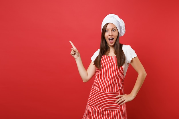 Huisvrouw vrouwelijke chef-kok of bakker in gestreepte schort, wit t-shirt, toque chef-koks hoed geïsoleerd op rode muur achtergrond. Mooie huishoudster vrouw wijsvinger omhoog. Mock-up kopie ruimteconcept.