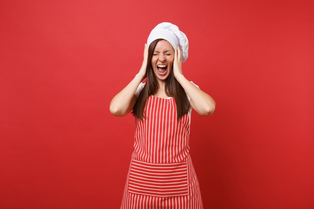 Huisvrouw vrouwelijke chef-kok of bakker in gestreepte schort, wit t-shirt, toque chef-koks hoed geïsoleerd op rode muur achtergrond. Schreeuwende geschokte huishoudstervrouw legde handen op het hoofd. Mock-up kopie ruimteconcept.