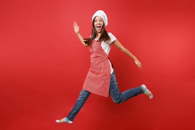 Huisvrouw vrouwelijke chef-kok of bakker in gestreepte schort wit t-shirt, toque chef-koks hoed geïsoleerd op rode muur achtergrond. Volledige lengte portret huishoudster vrouw hoog springen. Bespotten kopie ruimte concept.