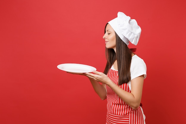 Huisvrouw vrouwelijke chef-kok of bakker in gestreepte schort, wit t-shirt, toque chef-koks hoed geïsoleerd op rode muur achtergrond. Vrouw houdt lege lege ronde plaat met plaats voor voedsel. Mock-up kopie ruimte concept