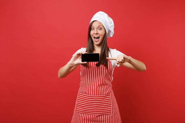 Huisvrouw vrouwelijke chef-kok of bakker in gestreepte schort wit t-shirt toque chef-koks hoed geïsoleerd op rode muur achtergrond. Vrouw houdt mobiel leeg scherm voor promotionele inhoud Mock-up kopie ruimteconcept