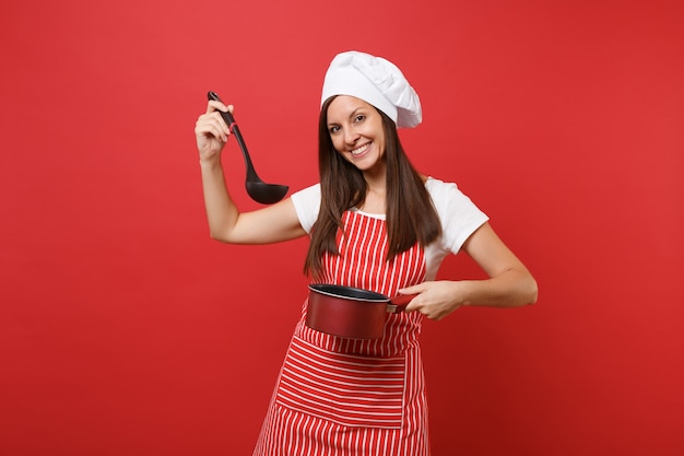 Huisvrouw vrouwelijke chef-kok of bakker in gestreepte schort wit t-shirt toque chef-koks hoed geïsoleerd op rode muur achtergrond. Vrouw houdt proeverij lege kookpan zwarte soeplepel dipper. Mock-up kopie ruimte concept
